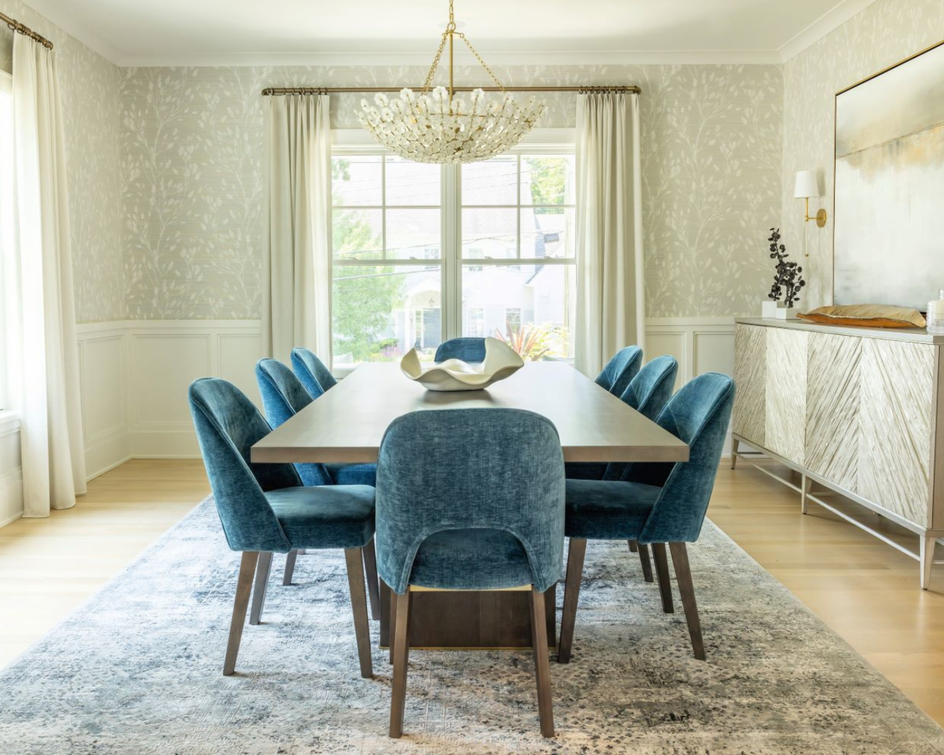 Photo of bright modern dining room with white walls, long dining room tables surrounded by velvet turquoise chairs, and large crystal chandelier hanging above table.