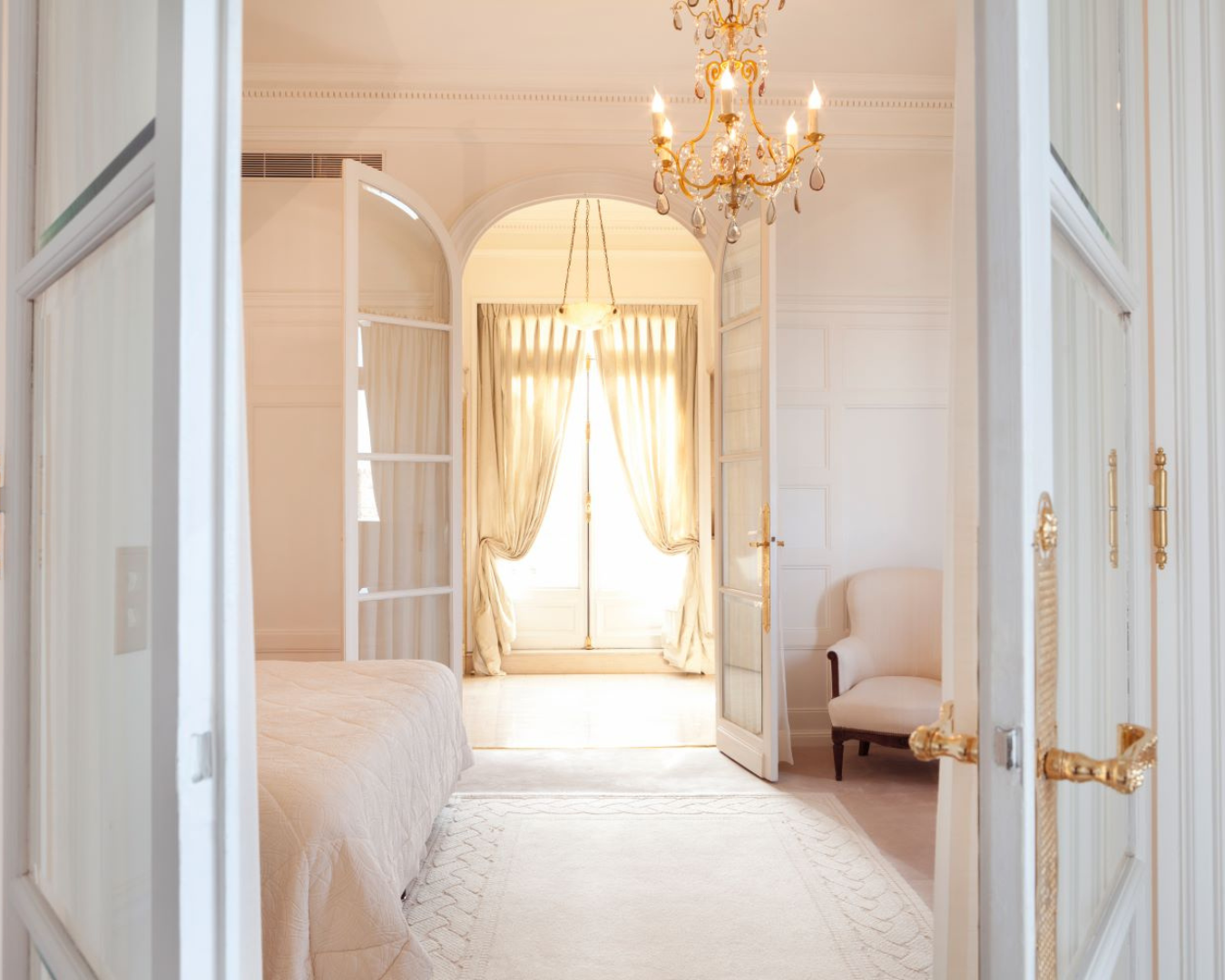 Photo from Castilian Collection. Beautiful, large bedroom with white and light pink accents, hold chandelier, and large window with gold drapery hardware, paired with delicate, sheer drapes.